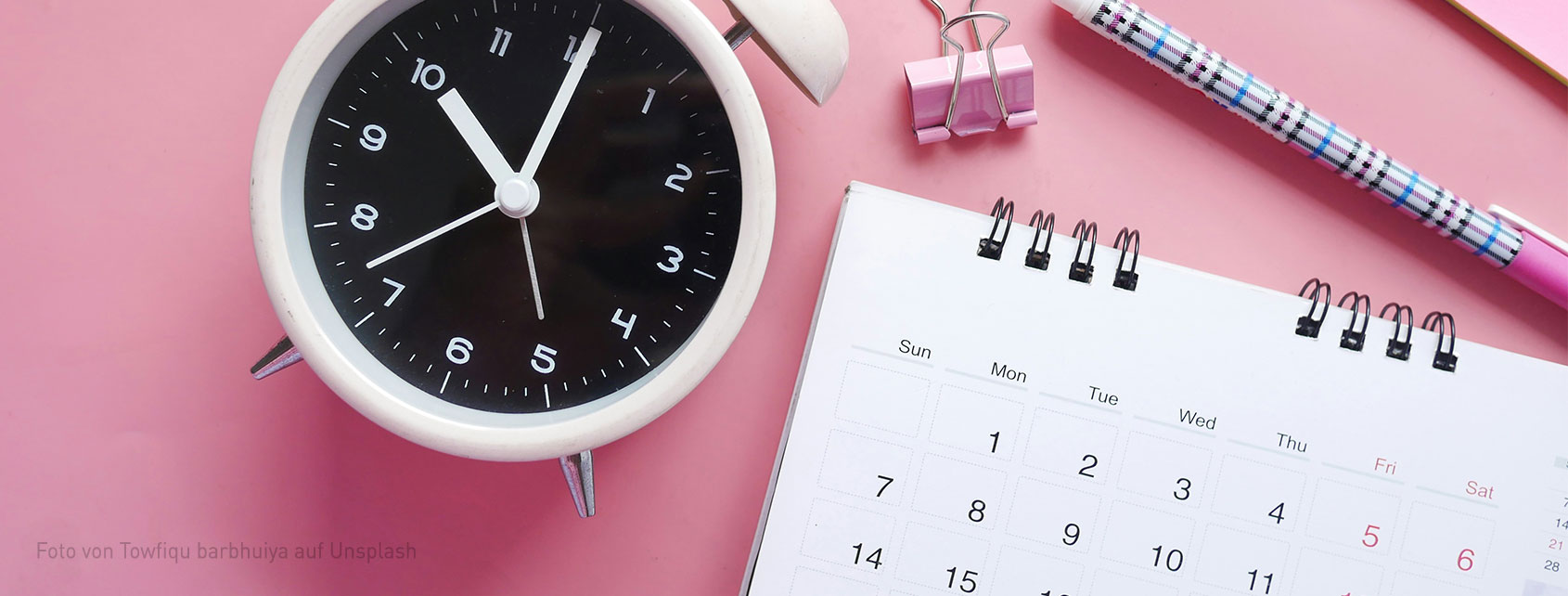 Ein Wecker neben einem aufgeschlagenen Tischkalender neben Stiften auf rosa Untergrund.